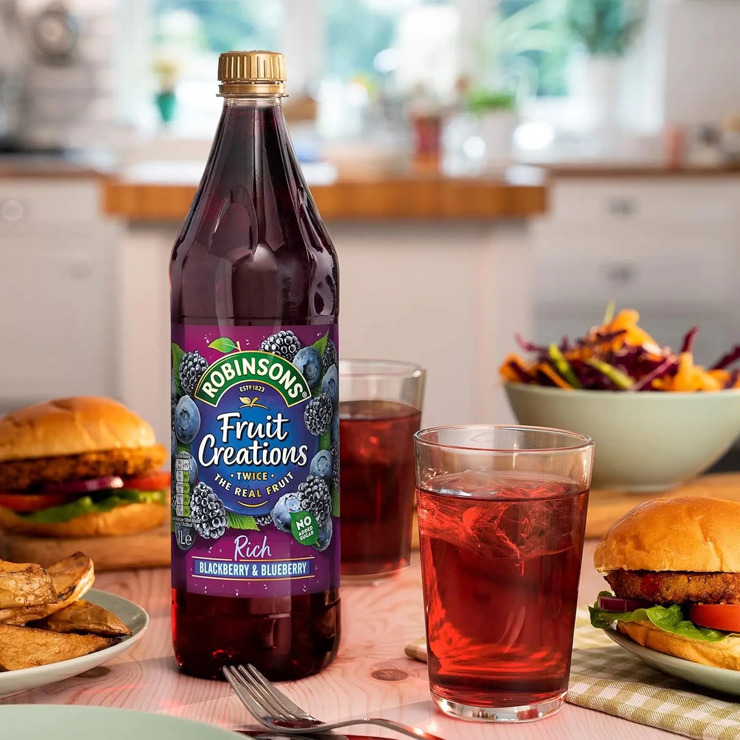Bottle of Robinsons Fruit Creations Blackberry & Blueberry juice on a table with burgers and a salad in a kitchen setting.