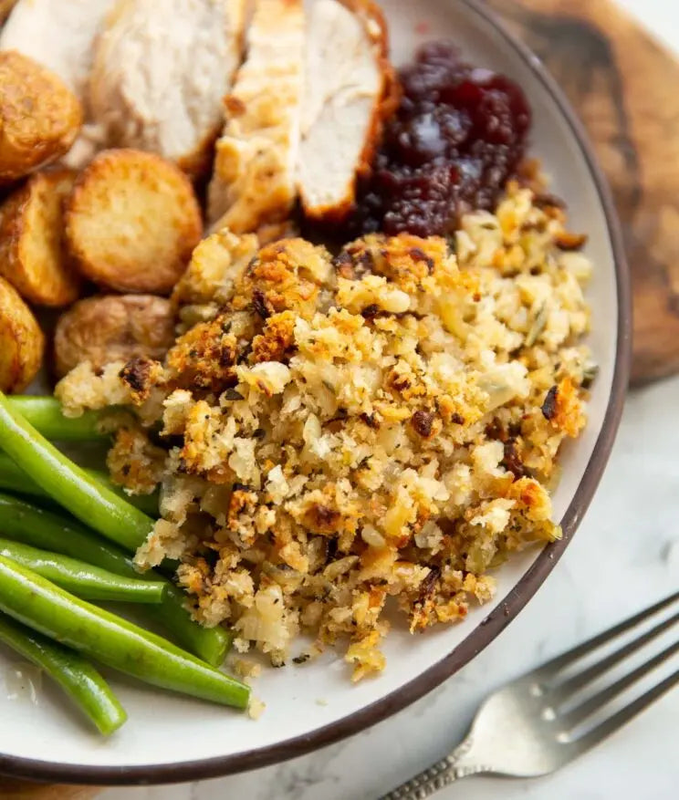 A close-up of a plated Thanksgiving meal featuring stuffing, sliced turkey, green beans, roasted potatoes, and cranberry sauce on a white plate with a dark rim.