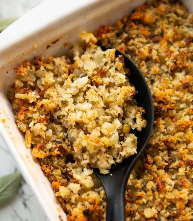 A close-up shot of a creamy stuffing or dressing in a white ceramic baking dish, with a black serving spoon lifting a portion. The stuffing has a golden-brown, crumbly texture with visible herbs and seeds.