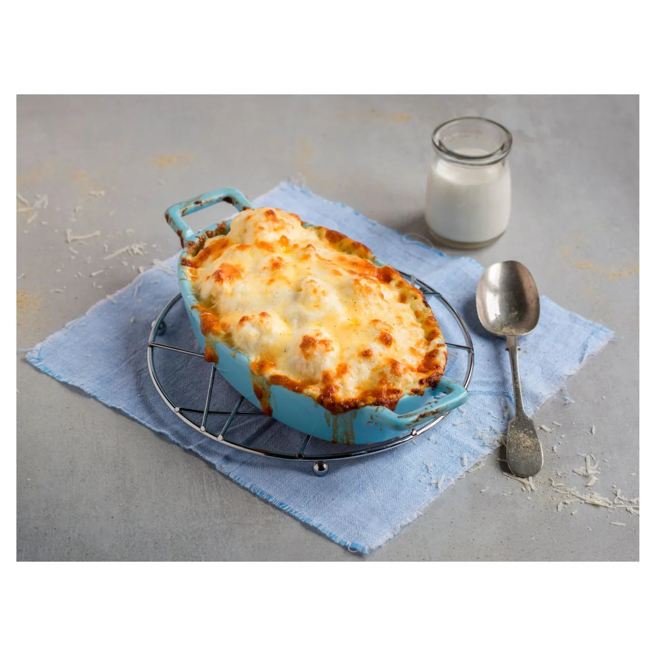 Cheesy dish in a blue baking dish on a gray surface with a glass of milk and spoon.