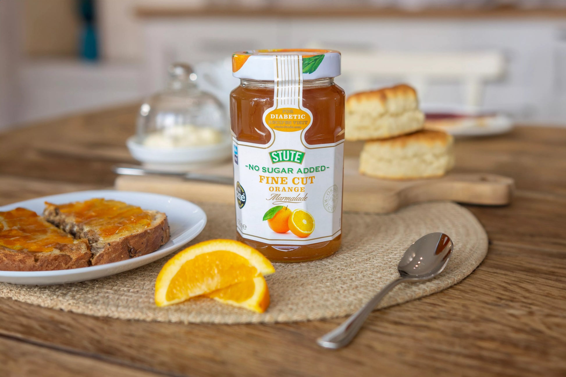Jar of Stute no sugar added fine cut orange jam on a wooden table with bread and orange slices.