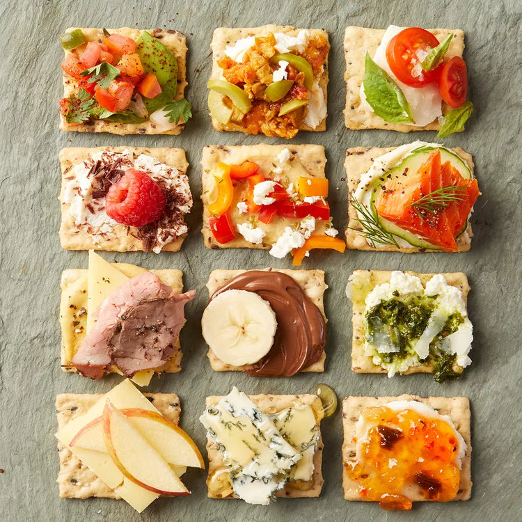 A grid of twelve assorted finger crackers topped with various colorful foods, including tomatoes, cheese, fruit, meats, and spreads, arranged on a textured gray slate background.