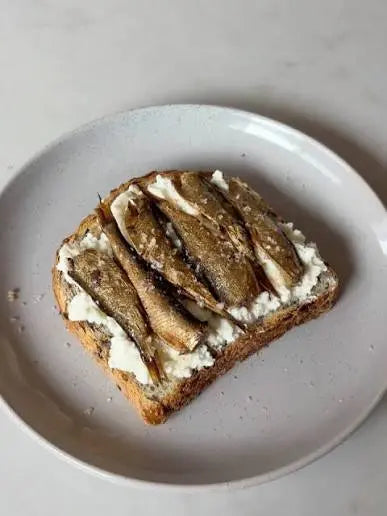 A close-up shot of a slice of toasted bread spread with white cheese and topped with several small grilled or fried fish, served on a light gray ceramic plate against a neutral light gray background.