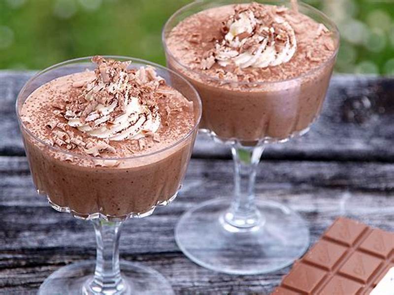 Two glasses of chocolate mousse with whipped cream on a wooden surface, with a bar of chocolate in the foreground.